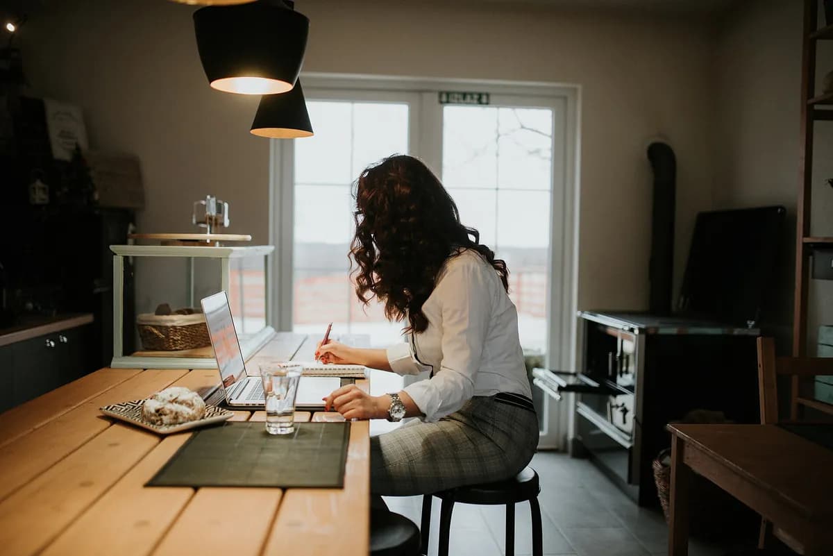 Woman writing notes beside her laptop while working at a wooden table