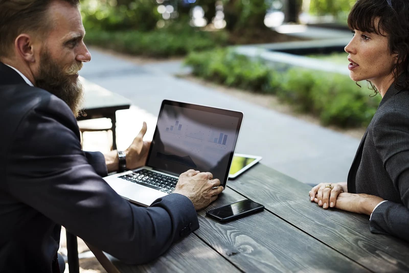 Two professionals in business attire talking at an outdoor table with a laptop